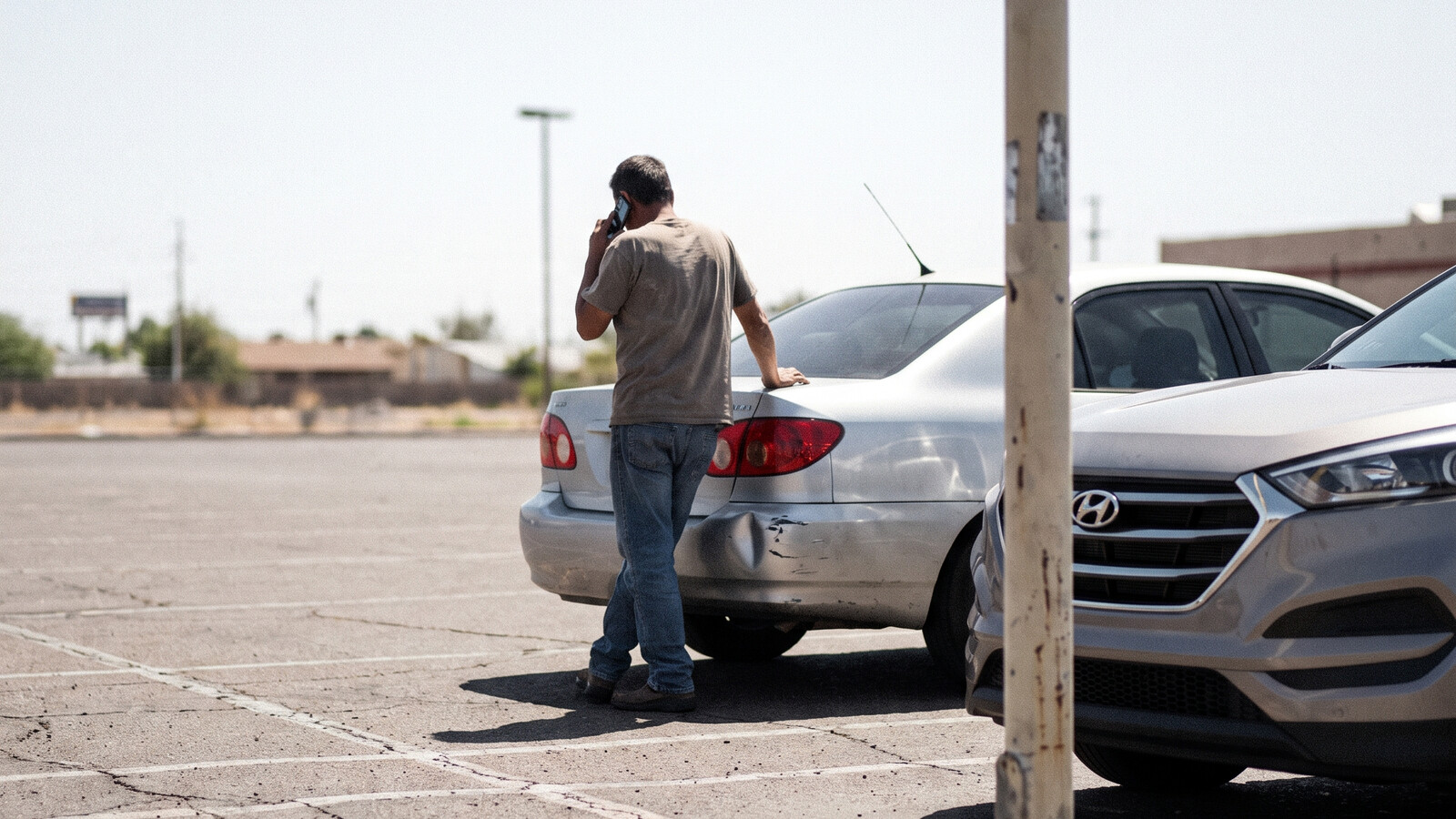 A man on the phone beside his dented car in a Phoenix parking lot after a crash